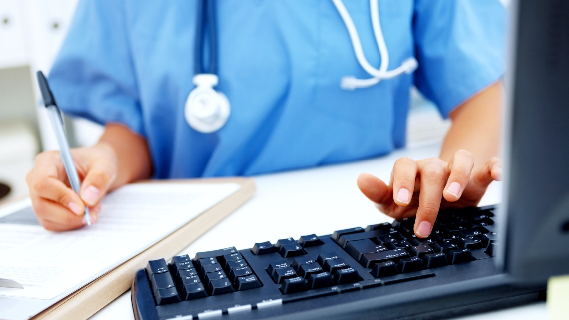 A nurse works at a desk with a computer and a clipboard