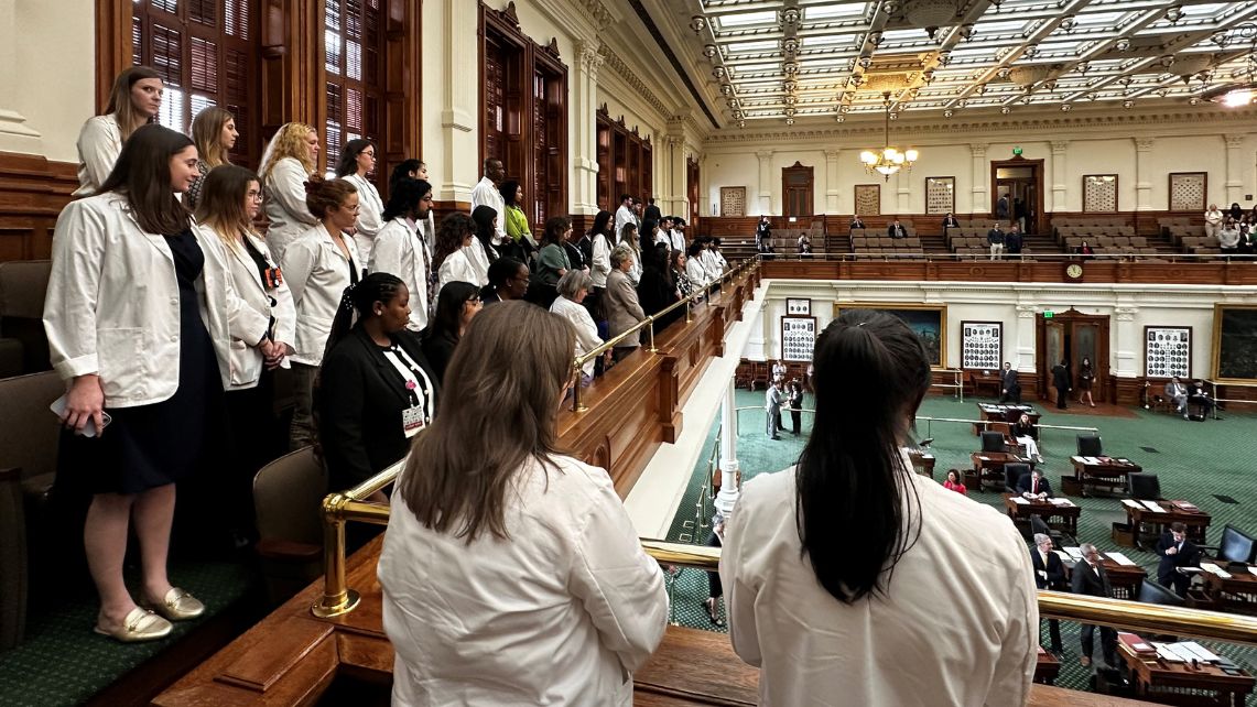 UTMB students in white coats stand in the gallery of the Texas Senate as they are recognized by the senators on the floor