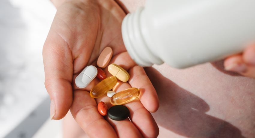 A person holds a handful of multicolored pills and capsules in one hand, representing the many nutritional supplements people may take. The image illustrates the importance of understanding what supplements are needed and how they may affect health.