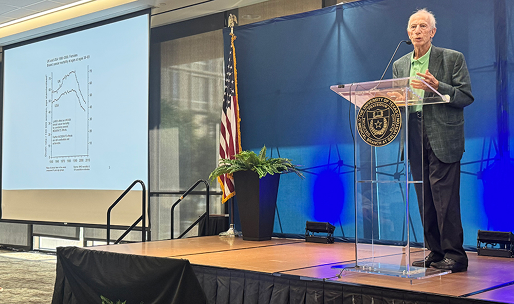 A speaker stands at a podium bearing the UTMB seal in Levin Hall, presenting research during the “Navigating Dual Frontiers: Psychiatry & Women’s Health Symposium,” with a slide displaying data on a large screen and an American flag on stage