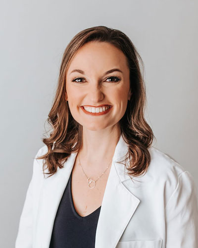Professional headshot of a person wearing a white coat over a dark top, facing the camera against a light gray background.