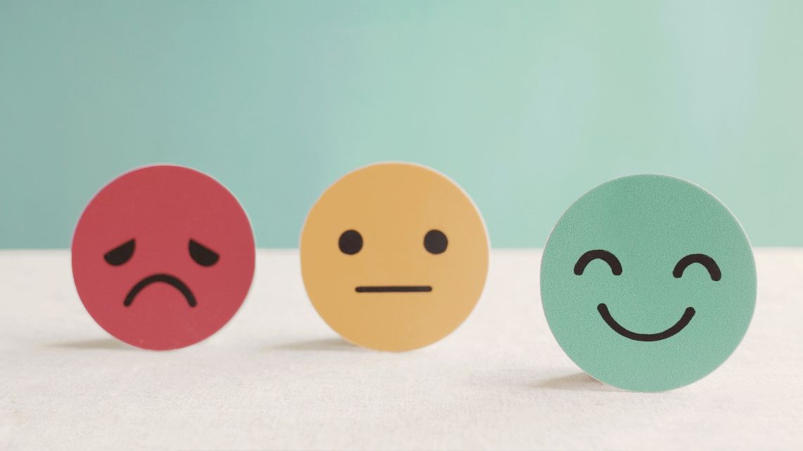 Three round icons sit side by side on a light surface, each showing a different facial expression: a red frowning face, a yellow neutral face, and a green smiling face against a teal background