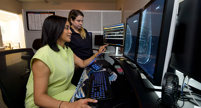 Two people seated at a workstation review breast imaging scans displayed on multiple computer monitors, with one person pointing to a screen.