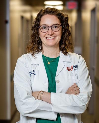 Person in a white clinical coat stands in a hallway with arms crossed. A UTMB Health logo is visible on the coat.