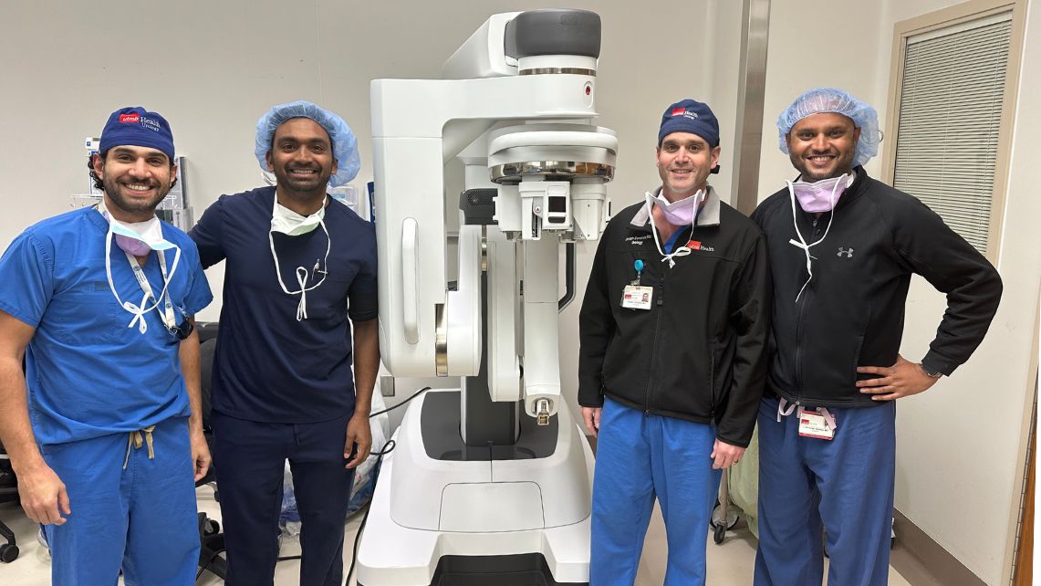 Four medical professionals in blue scrubs stand around a robot