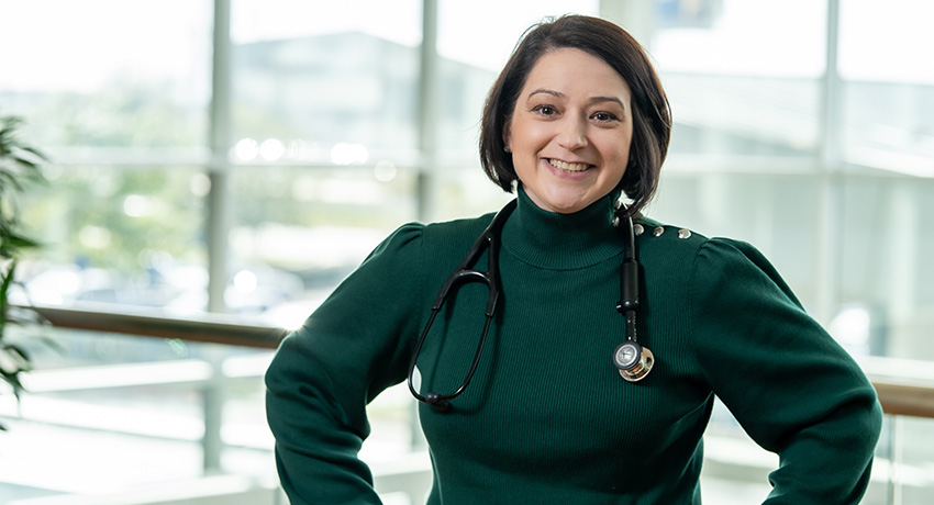 A woman wearing a green sweater and stethoscope stands in a bright hospital atrium.