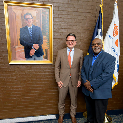 Two people stand in front of a framed portrait and two flags against a brick wall. One wears a tan suit with an orange tie, and the other wears a blue shirt under a dark jacket.