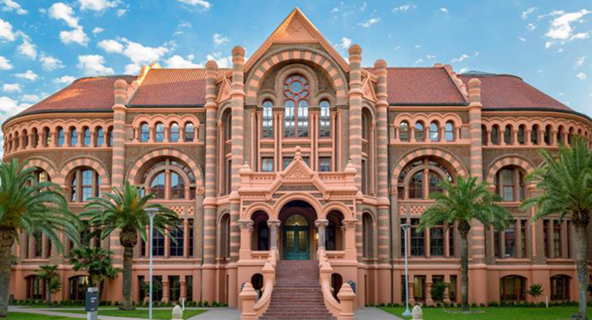 Exterior view of UTMB’s historic campus building with red‑brick Romanesque architecture, arched windows, palm trees, and a central staircase under a partly cloudy sky