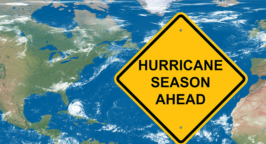 Yellow diamond-shaped warning sign reading “Hurricane Season Ahead” overlaid on a satellite map of the Atlantic Ocean, with the southeastern United States, Caribbean, and a visible storm system swirling over the water.