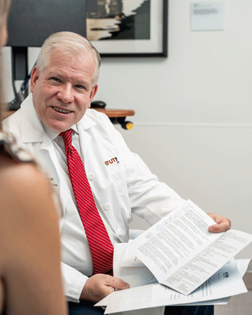Person in a white medical coat and red tie sits holding several printed documents while speaking with another seated person in an office setting.