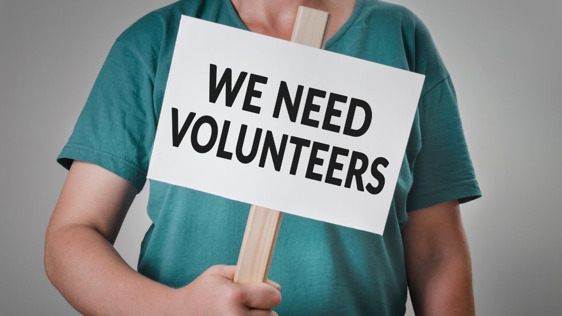 A man in green scrubs holds a sign on a wooden picket that reads "We need volunteers"