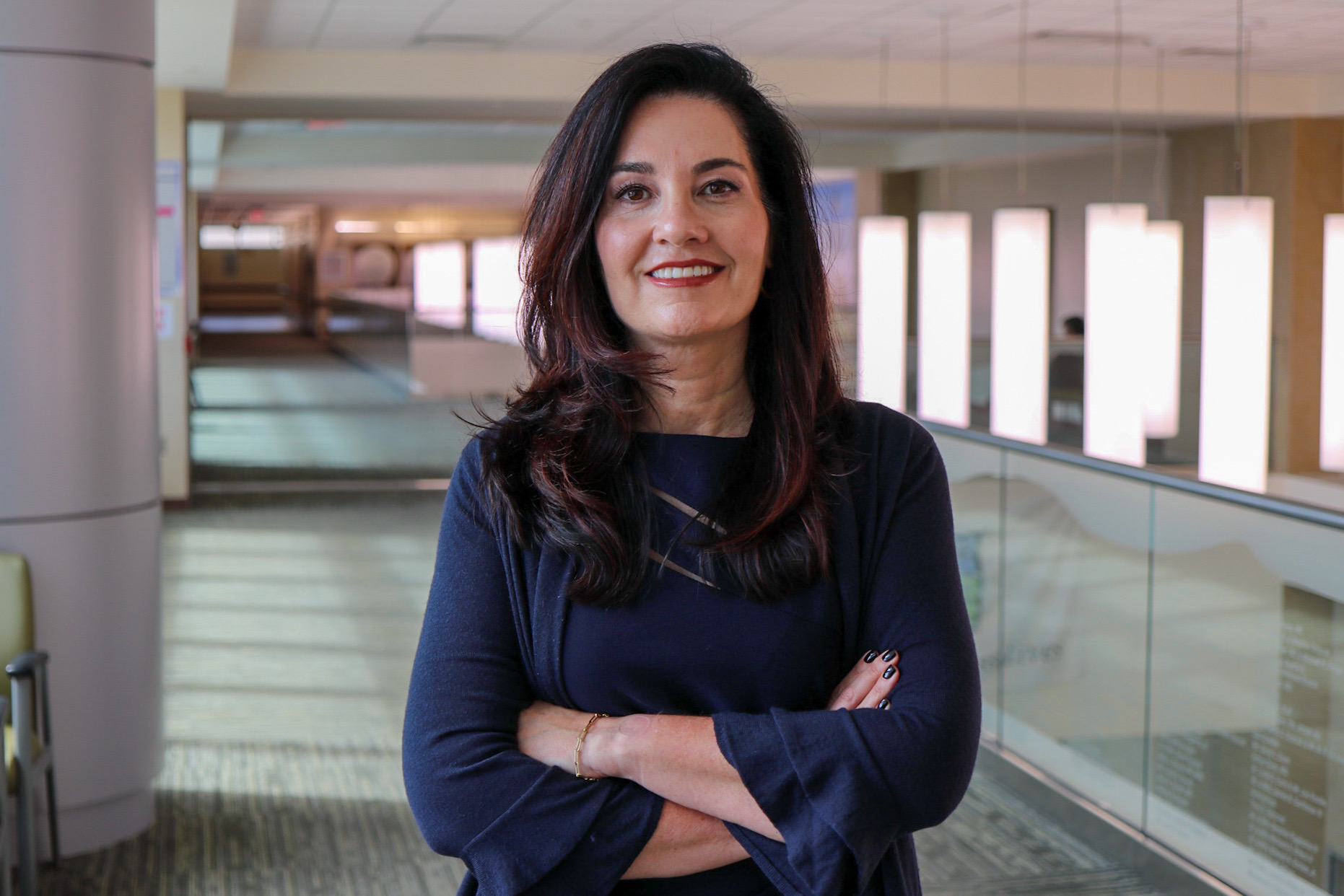 A person standing indoors with arms crossed in a hallway with bright rectangular lights and glass railing in the background.