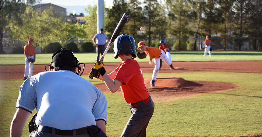 Youth baseball game in progress with a batter at home plate, catcher and umpire crouched behind, and a pitcher throwing the ball from the mound on a sunny outdoor field