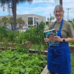 Standing by the OLLI garden, Allison Brown shows off her Blue Zone cookbook H A person wearing a blue apron stands in a raised garden bed area and holds a cookbook. Garden plants, trees, and a building are visible in the background.