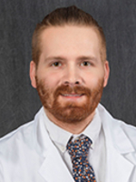 Head-and-shoulders studio portrait of a person wearing a white lab coat over a dress shirt and patterned tie, photographed against a textured dark gray background.