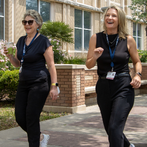 Two women walk side by side on a paved campus walkway, wearing black outfits and UTMB lanyards, with a brick building, windows, and landscaping in the background.