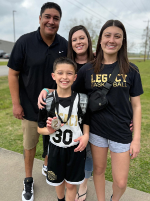 Four people stand together outdoors on a sidewalk; a child in a sports jersey and backpack holds a medal in the foreground, with three other individuals behind on a grassy area. 