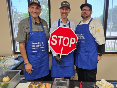 From left: Craig Kovacevich, GISD Superintendent Matthew Neighbors and Cheg Kelsey Thomas prepare to make Guacamole & Bean Tacos. Three people wearing blue UTMB aprons stand behind a table and hold a red octagonal STOP sign. Cooking supplies and food preparation materials are visible on the table.