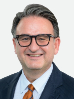  Professional head-and-shoulders photo shows a man with short dark hair and glasses wearing a dark blue suit and orange tie with a light blue shirt. 