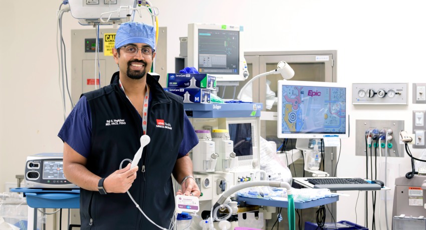 A surgeon in scrubs and a UTMB vest stands in an operating room, smiling while holding a handheld medical device, with monitors and surgical equipment visible in the background.
