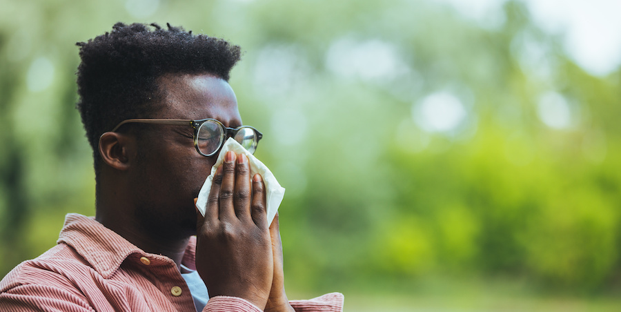 image of black man sneezing and blowing nose into tissue - in the background is blurred out greenery. The man is wearing glasses and a collared shirt