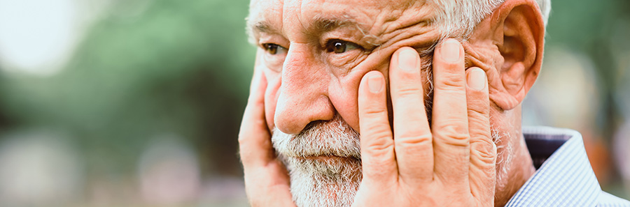 Close-up of an older adult resting both hands against the face while looking downward outdoors, with a softly blurred green background