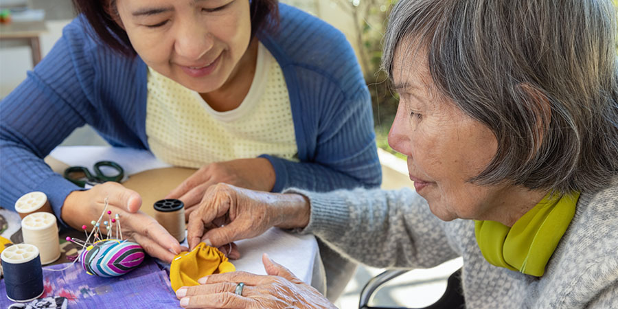 Caregiver and older adult sit at a table working on a small sewing project, with spools of thread, pins, and fabric laid out, illustrating hands‑on support and daily activities for aging adults