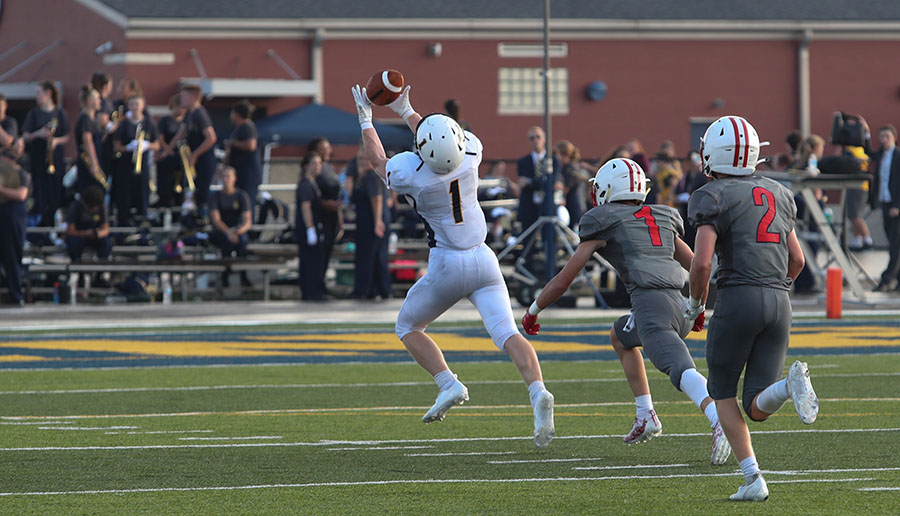 Youth football players compete on a field during a game, with one player leaping to catch a pass while two defenders pursue, as spectators watch from the bleachers in the background