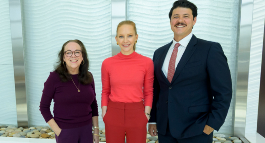 Two women and a man are shown standing in front of a white textured wall, with decorative stones visible at the base of the wall. One person wears purple, one wears a bright coral top and matching pants, and one wears a navy suit with a red tie.