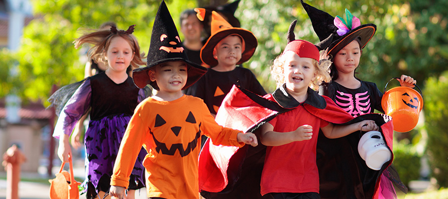 Group of children wearing Halloween costumes walk together along a residential sidewalk, holding trick‑or‑treat buckets, with an adult visible in the background and trees lining the street