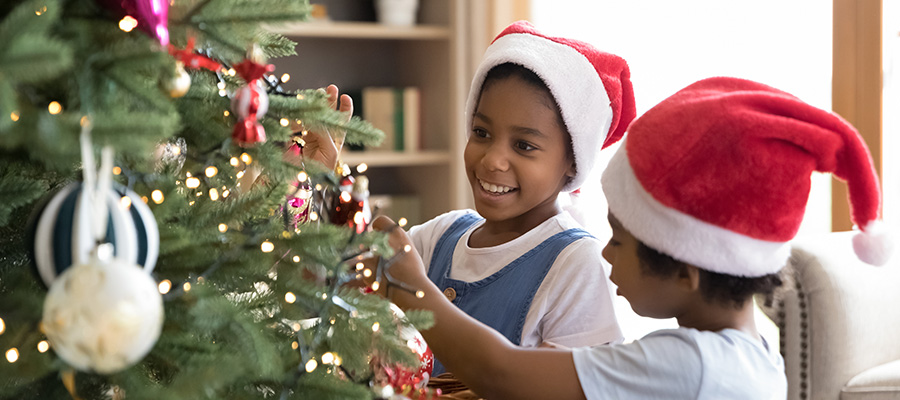 Two children wearing Santa hats decorate a Christmas tree with ornaments and lights inside a living room