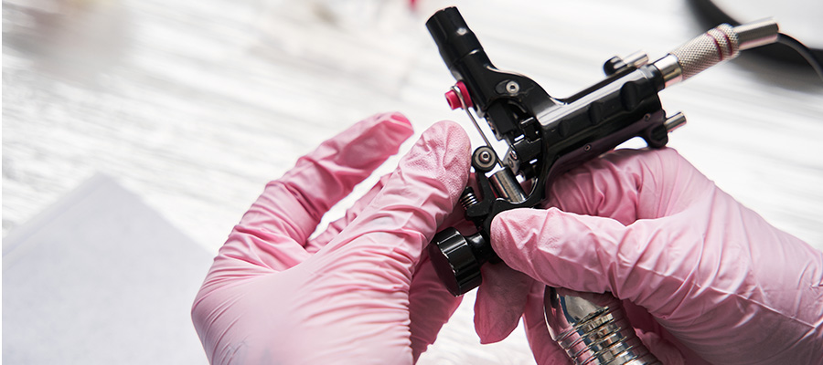 Close‑up of gloved hands holding a tattoo machine over a clean work surface, illustrating a medical tattooing procedure used in breast reconstruction care