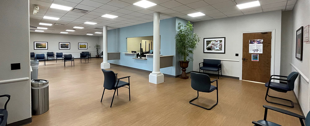 Renovated Regional Women’s Services & Pediatrics clinic lobby with light blue walls, wood‑tone floors, spaced seating, framed artwork, indoor plants, and a central reception window creating a bright, welcoming space for patients and families