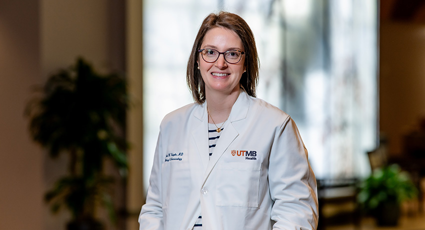 A woman wearing glasses and a white UTMB Health lab coat is standing indoors against a softly blurred background. A plant and large windows are visible behind the person.