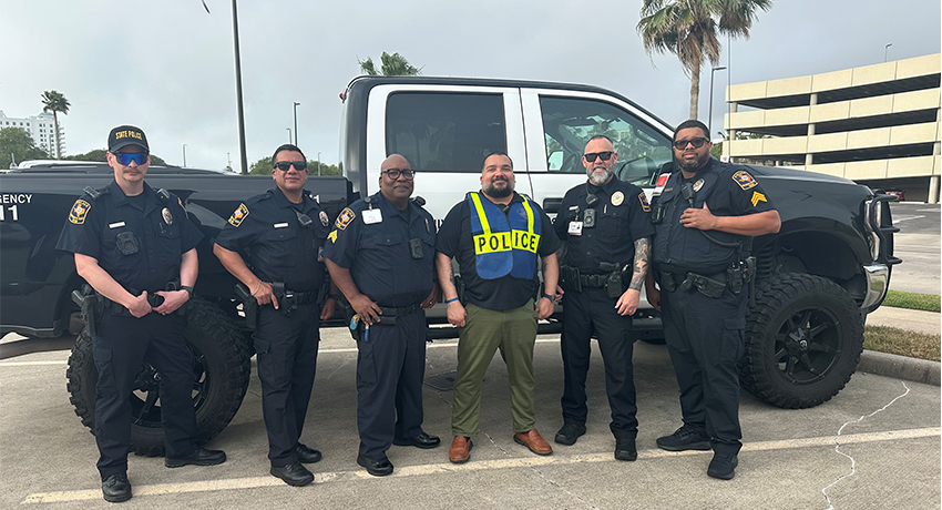Six uniformed UTMB police officers stand in a parking lot in front of a police truck. One officer wears a high‑visibility vest with the word “POLICE” printed on the front.
