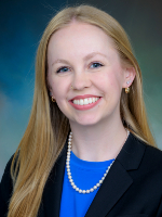 Head-and-shoulders shot of woman with blonde hair, blue shirt and black jacket on a studio blue background” 